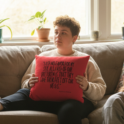 A queer person in a cream sweater sits on a gray couch, holding the “No Favor For My Sex Pillow” with black handwritten text.