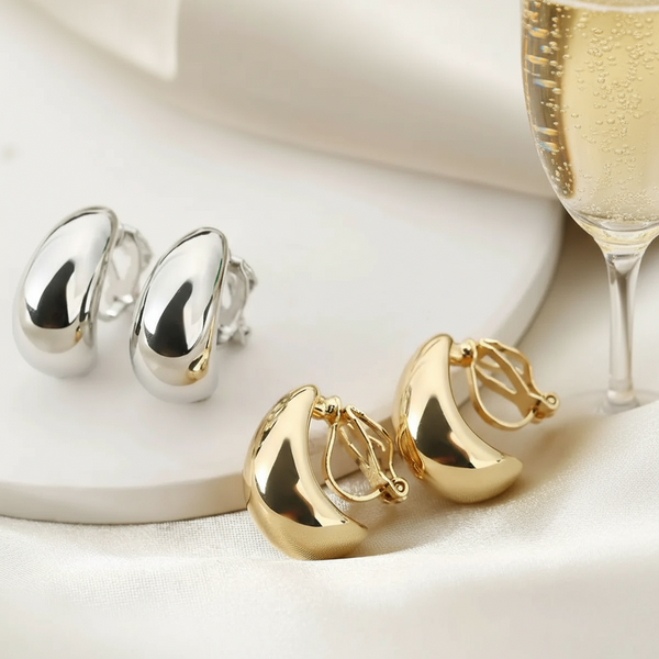 Silver and gold crescent-shaped clip-on earrings displayed beside a glass of champagne