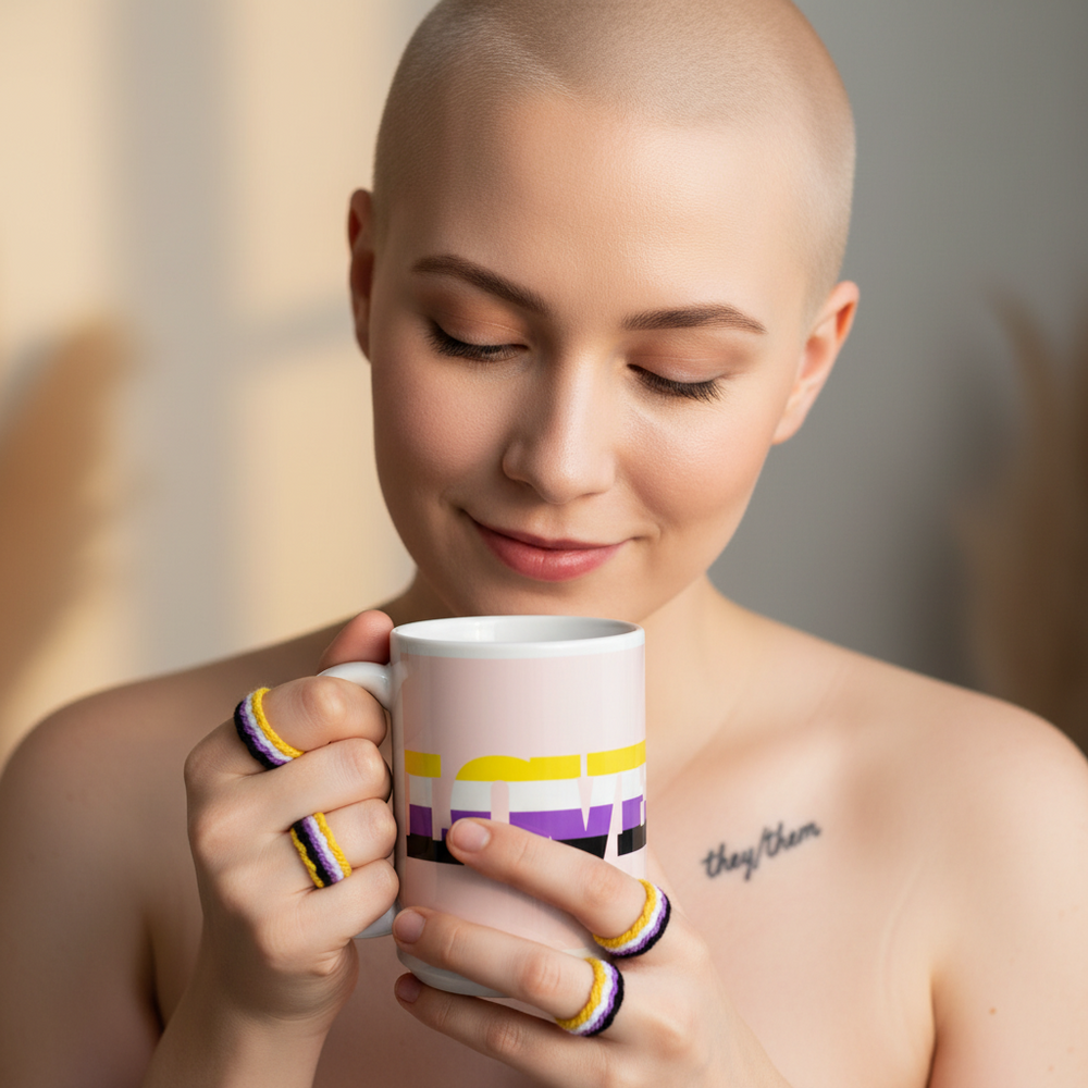 Non-Binary Love Mug held by a bald, person with purple and yellow rings, “they/them” collarbone tattoo, soft lighting