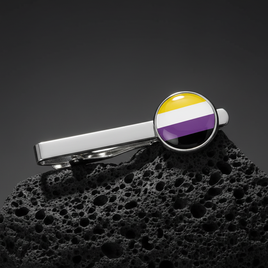 Non-Binary Pride tie clip with a round yellow, white, purple, and black flag emblem on silver, shown on black volcanic rock.