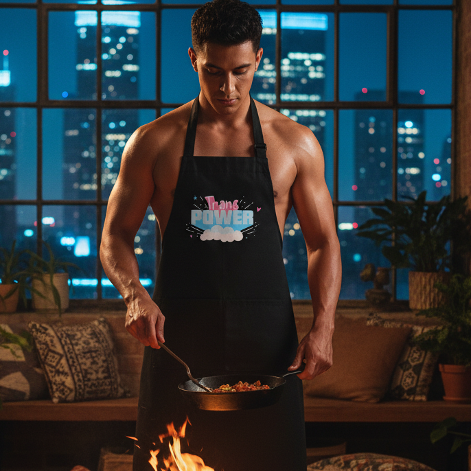 A young man cooks in a loft, wearing the Stand Proud Organic Cotton Apron with Trans Power text, embodying bold, sexy queer pride and self-expression.