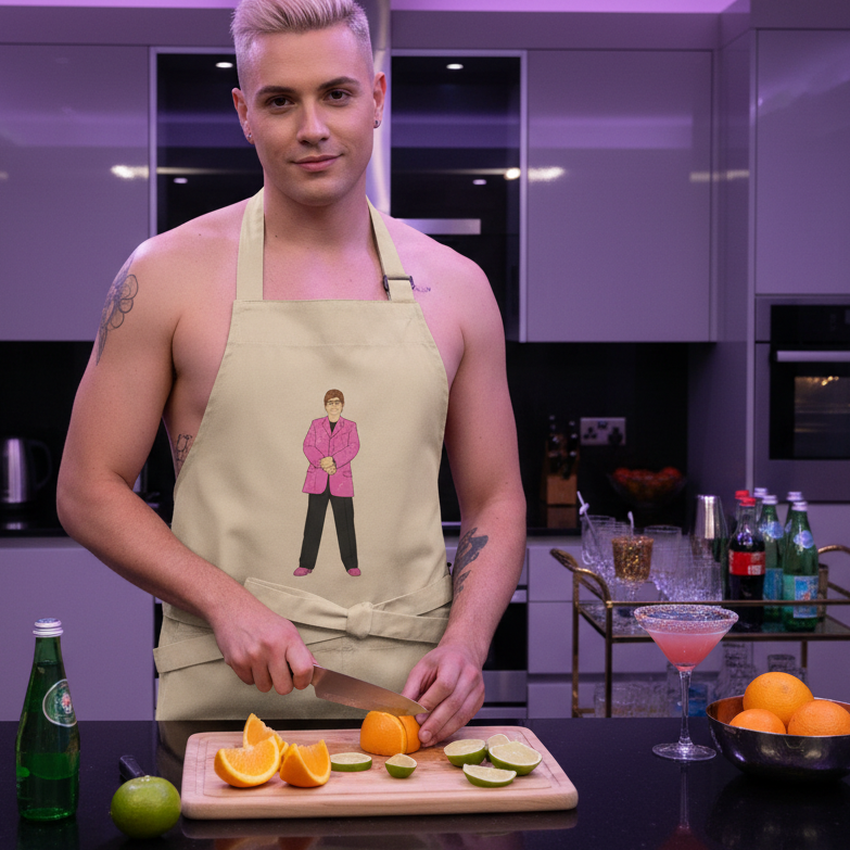 Young man in a sleek kitchen wears the bold Elton John Organic Cotton Apron with a cartoon figure, slicing citrus fruits, embodying pride and self-expression amidst vibrant, queer kitchen vibes.