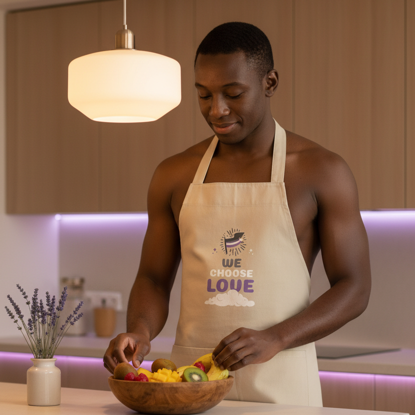 Young man in a kitchen wears the Asexual We Choose Love Organic Cotton Apron, adorned with a stylized bird and positive message, arranging fruit in a bowl, embodying pride, confidence, and self-expression.