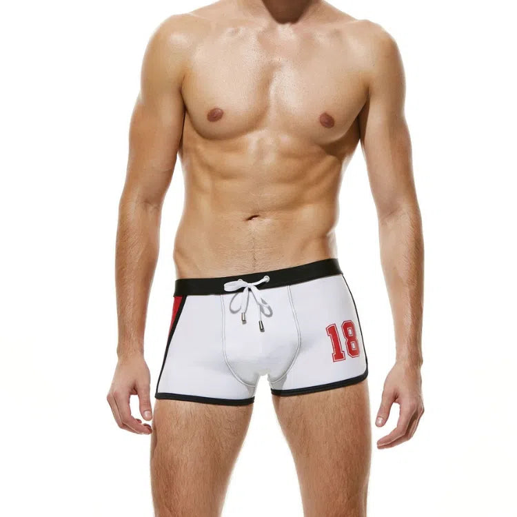 Front-facing model wearing white boxer-style swim trunks with black trim and a red 18