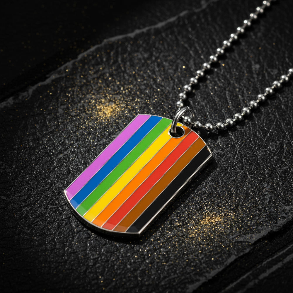 Rainbow-striped rectangular tag pendant on a ball chain, lying on black fabric with gold flecks