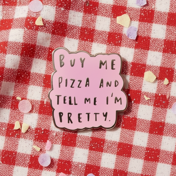 Pink enamel pin reading “Buy Me Pizza and Tell Me I'm Pretty” on red gingham fabric