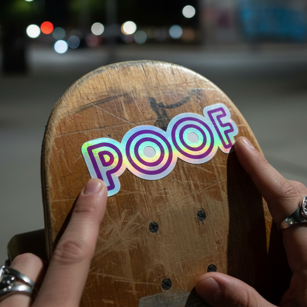 A Poof holographic sticker is pressed onto a scuffed skateboard deck at night by ring-adorned hands