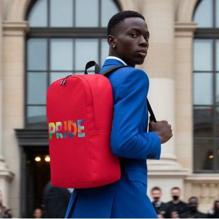 Pride Minimalist Backpack with bold, colorful LGBTQ+ letters, worn by someone in a bright blue suit outside a grand building.