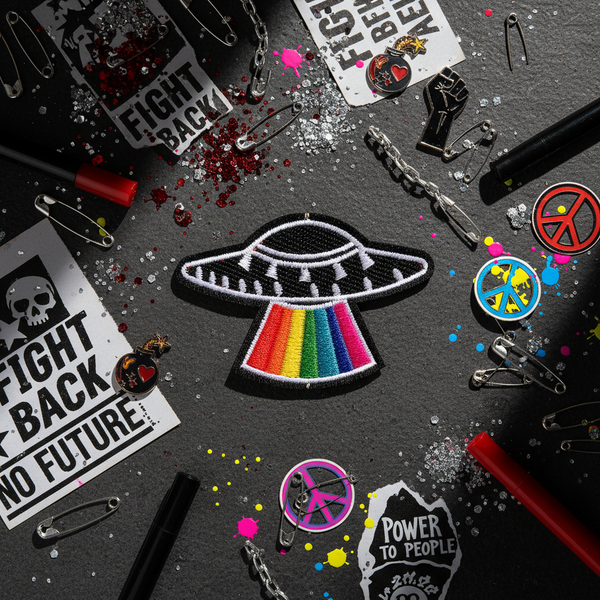 Pride rainbow UFO iron-on patch on dark background with protest stickers, peace signs, paint splatters, red pens, pins