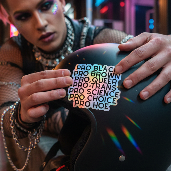 Pro Hoe (Black Text) holographic sticker shines with rainbow vibes as a queer person in glitter places it on a black helmet.