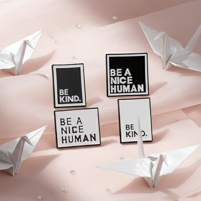 Flat-lay of four black-and-white 'Be A Nice Human' enamel pins on a pink satin background, surrounded by origami cranes and crystal beads, embodying queer pride and self-expression.