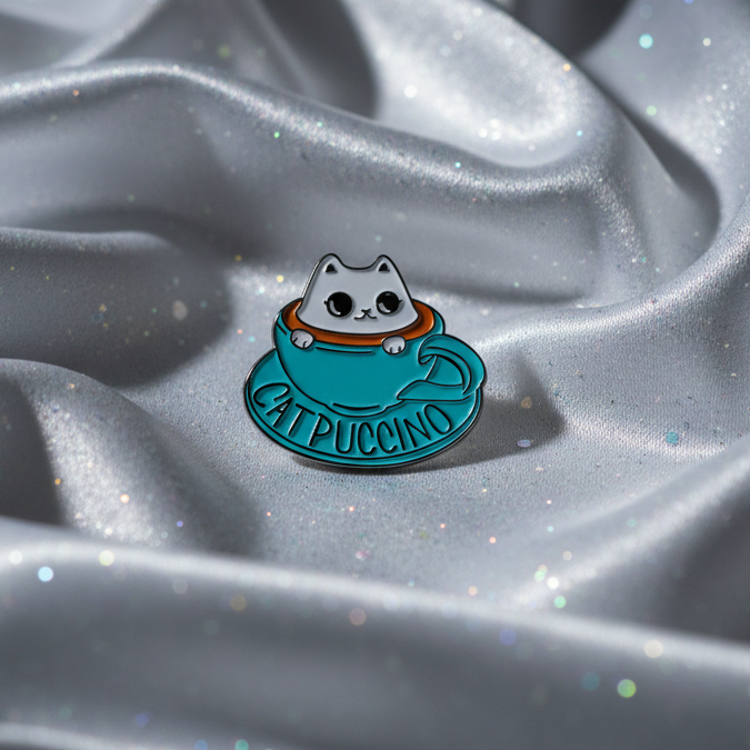 Close-up of a cat-shaped Catpuccino enamel pin on a turquoise coffee cup
