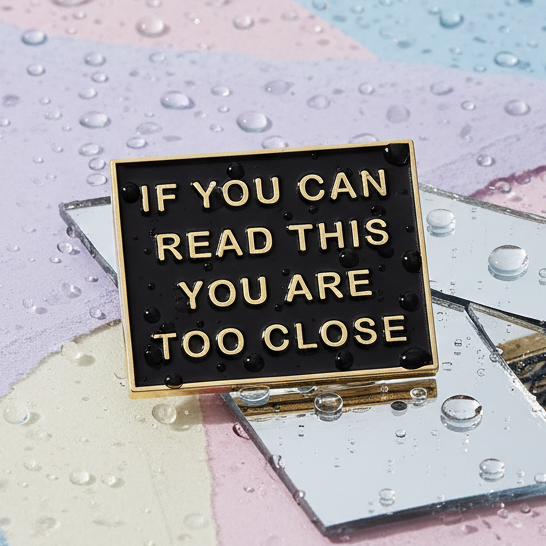 If You Can Read This You Are Too Close Enamel Pin: Black-and-gold pin on mirror shards with water droplets. Bold queer statement, perfect for expressing fierce pride and visibility.