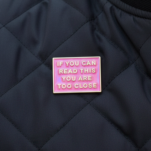 Pink enamel pin reading “IF YOU CAN READ THIS YOU ARE TOO CLOSE” on black quilted fabric