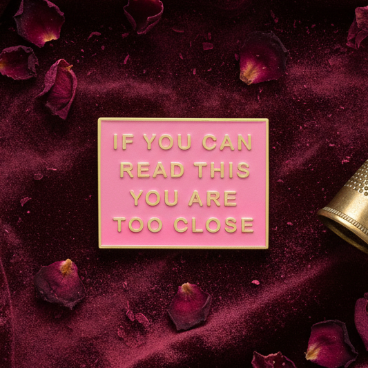 If You Can Read This You Are Too Close Enamel Pin on plush fabric, surrounded by rose petals. The pin's bold gold lettering on pink embodies fierce, proud queer self-expression and empowerment.