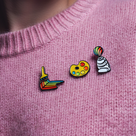 Close-up of Rainbow Paint Brush Enamel Pins on a pink knit sweater, featuring a rainbow-bristled brush, a vibrant artist's palette, and a striped paint tube, celebrating queer creativity and pride.