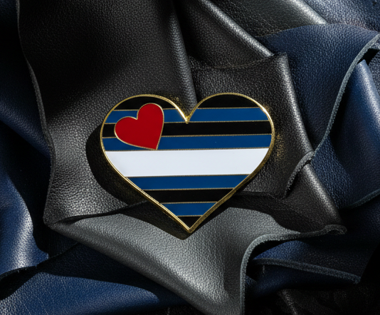 Heart-shaped Leather Pride Heart Enamel Pin on crumpled leather backdrop, featuring black, blue, and white stripes with a red heart. Celebrating LGBTQIA+ pride and self-expression with empowering style.