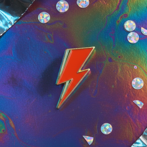 David Bowie Lightning Enamel Pin: A striking red and gold lightning-bolt pin on an iridescent backdrop, surrounded by holographic sequins. Celebrate queer pride and Bowie-inspired self-expression with this empowering accessory.