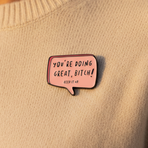 You're Doing Great Bitch Enamel Pin on a light-beige knitted sweater, features a pink speech bubble with an empowering message, embodying bold queer energy and prideful self-expression from Queer In The World: The Shop.
