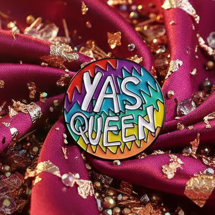 Yas Queen Enamel Pin on magenta-red fabric with gold accents, featuring a bold rainbow-gradient and white letters, embodying fierce LGBTQIA+ pride and empowerment.