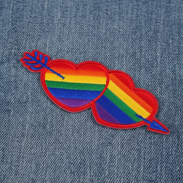 Rainbow Pride embroidered patch showing two overlapping hearts pierced by an arrow on denim