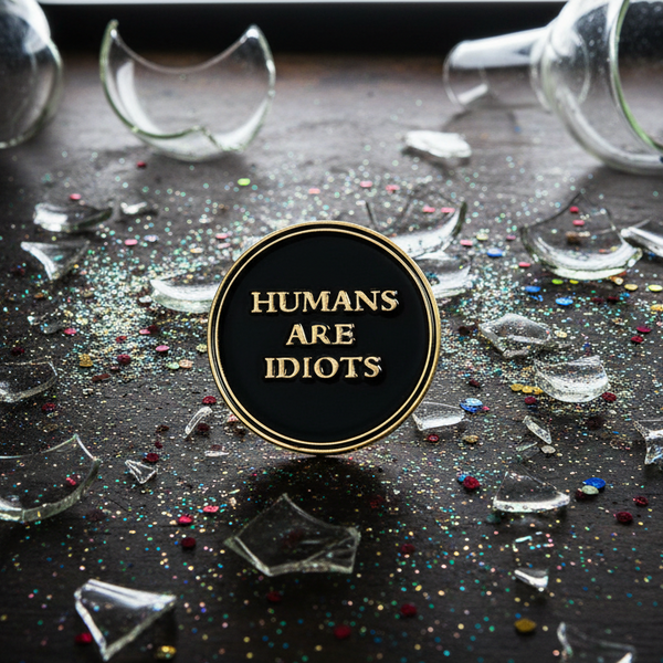 Black enamel pin with gold border and the text “HUMANS ARE IDIOTS,” amid broken glass and glitter
