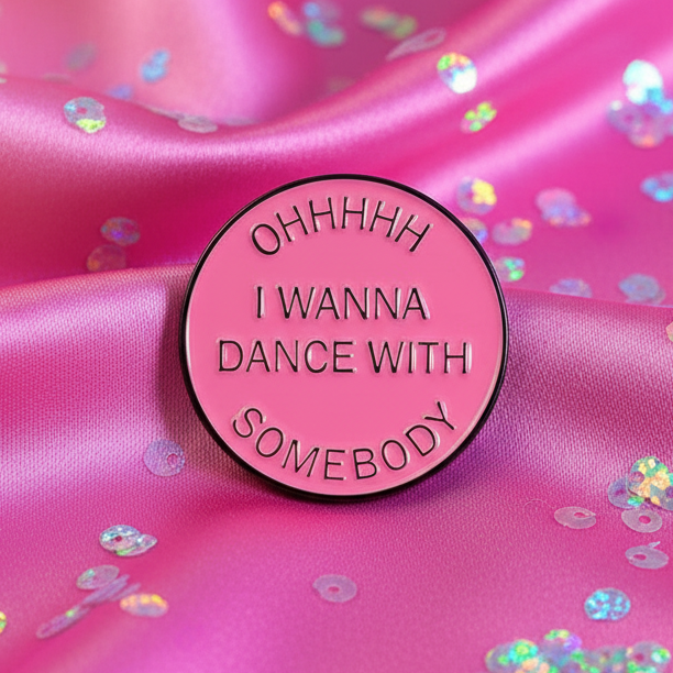 Round pink enamel pin embossed with OHHHHH I WANNA DANCE WITH SOMEBODY on satin-like fabric with iridescent confetti. An empowering symbol of pride and self-expression from Queer In The World.