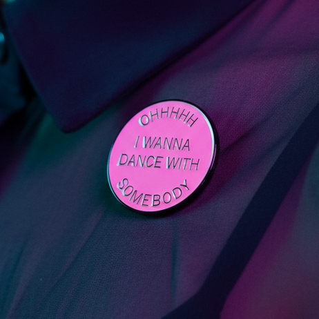 Pink enamel pin reading “OHHHHH I WANNA DANCE WITH SOMEBODY” on dark fabric