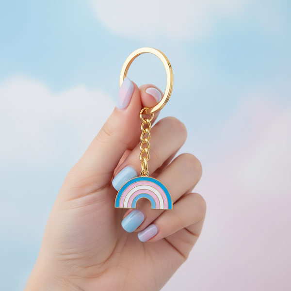 Hand holding a gold-tone keychain with a pink, blue, and white rainbow charm