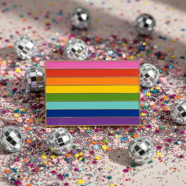 Close-up of a pink-striped LGBT Pride rainbow enamel pin amid confetti and disco balls