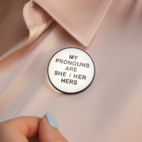 Close-up of a round enamel pin reading “MY PRONOUNS ARE SHE / HER HERS” on a shirt