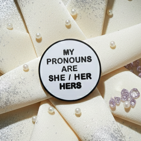 My Pronouns Are She/Her/Hers Enamel Pin with bold black lettering on a white background, radiates pride and visibility, resting on a textured surface adorned with faux pearls and glitter, embodying fierce self-expression.