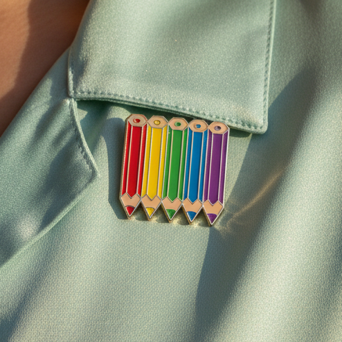 Rainbow-colored pencil enamel pin displayed on pale blue fabric