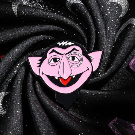 The Count Enamel Pin features a cartoon vampire head with a monocle, goatee, and fang, embodying queer pride and self-expression against a glittery black fabric with purple crystals.