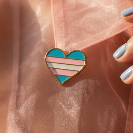 Trans Pride Heart Enamel Pin on peach fabric, outlined in gold and striped with light blue, pink, and white, symbolizing transgender pride. A hand with matching blue nails touches near the pin, embodying empowerment and self-expression.