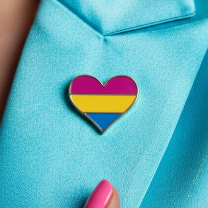 Pansexual Pride Heart Enamel Pin with pink, yellow, and blue stripes, pinned to a turquoise garment. A bold, pink-painted fingernail highlights this empowering, queer accessory, celebrating pride and self-expression.