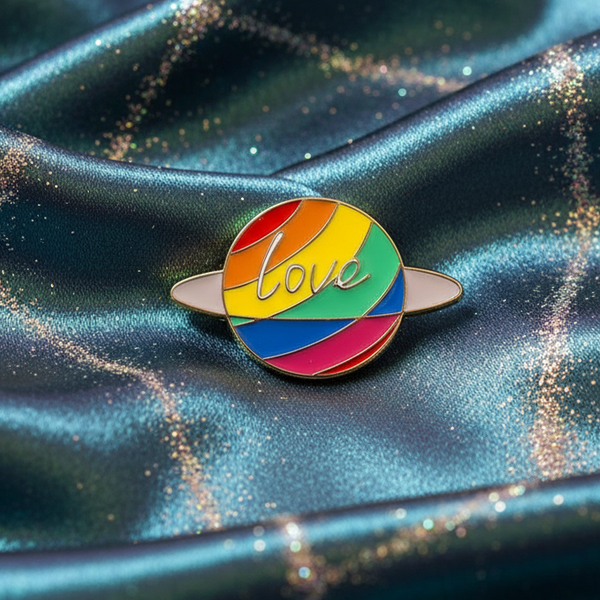Love Planet Enamel Pin featuring a rainbow-striped planet with rings and love in cursive, resting on shimmering teal-blue satin. Celebrate pride and self-expression with this empowering accessory.