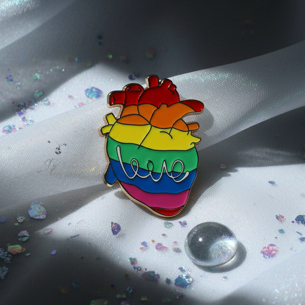 LGBT Heart Enamel Pin featuring a realistic heart with rainbow stripes and love script, symbolizing pride and empowerment, resting on glittered white fabric with a glass pebble.