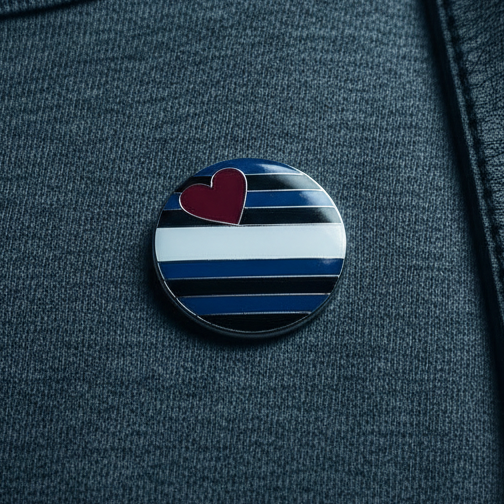 Close-up of a dark grey sweater displaying a Leather Pride Badge, featuring horizontal stripes and a red heart. A black leather strap crosses the chest, embodying bold, joyful queer pride and self-expression.