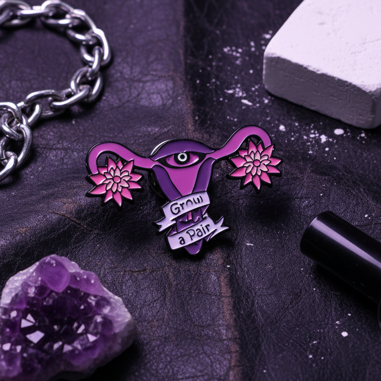 Purple enamel pin shaped like a flowering uterus with an eye and “Grow a Pair” banner