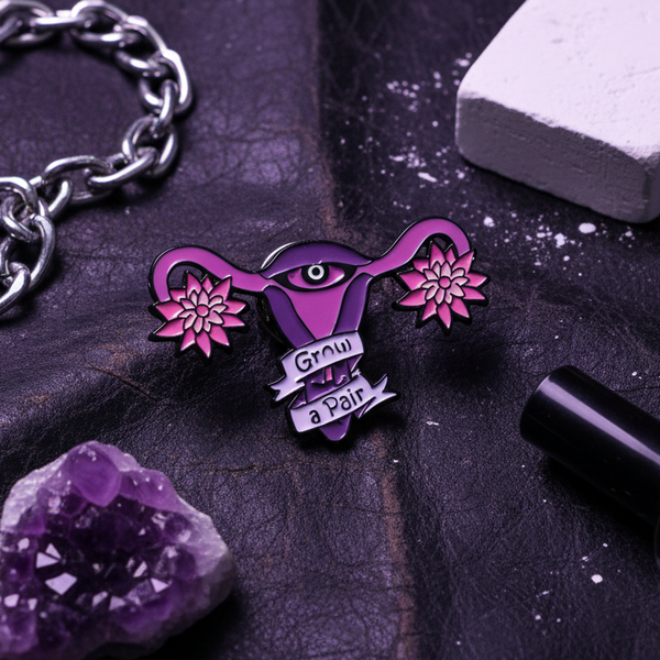 Purple enamel pin shaped like a flowering uterus with an eye and “Grow a Pair” banner