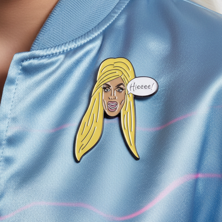 Blonde drag-queen face enamel pin with a “Hieeee!” speech bubble on blue fabric