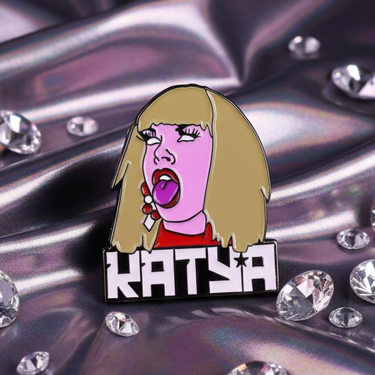Katya Enamel Pin features a vibrant cartoon portrait with blonde hair and bright pink skin, exuding playful, fierce energy. Perfect for celebrating pride and self-expression with bold visibility.