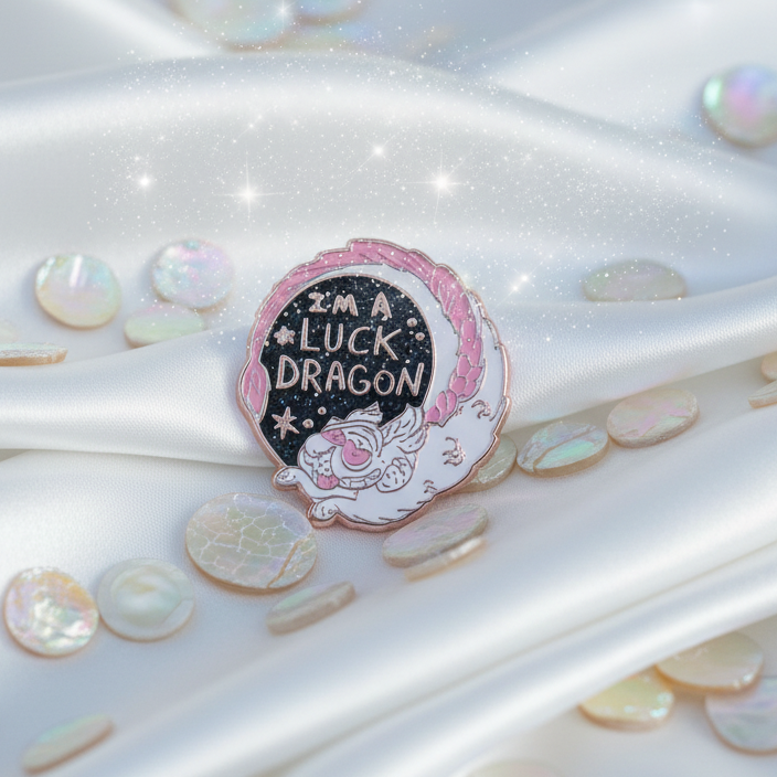 Close-up of the I'm A Luck Dragon Enamel Pin featuring a playful white and pink dragon on black enamel, accented with rose-gold. Set against satin and sequins, exuding queer pride and magic.
