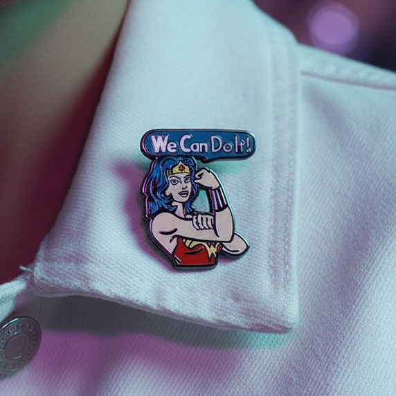 Close-up of a white denim jacket adorned with the We Can Do It! Enamel Pin, featuring Wonder Woman in a Rosie the Riveter pose, symbolizing empowerment and pride. A rainbow earring hints at vibrant self-expression.
