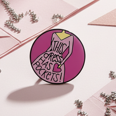 This Dress Has Pockets Enamel Pin features a pink stylized dress graphic with bold text. Set against a pink backdrop with metallic confetti, it embodies pride and self-expression.