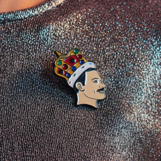 Freddie Mercury With A Crown Enamel Pin attached to a shiny metallic shirt; a bold, stylish accessory embodying queer pride and self-expression with a cartoon profile of a mustachioed man in a jeweled crown.