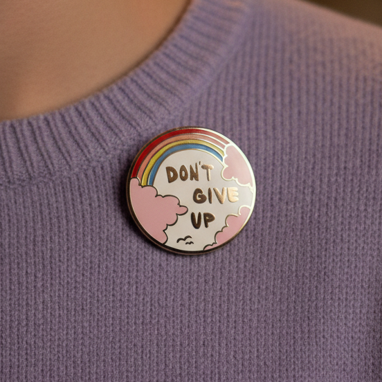 Rainbow enamel pin reading “DON’T GIVE UP,” with clouds on a purple sweater