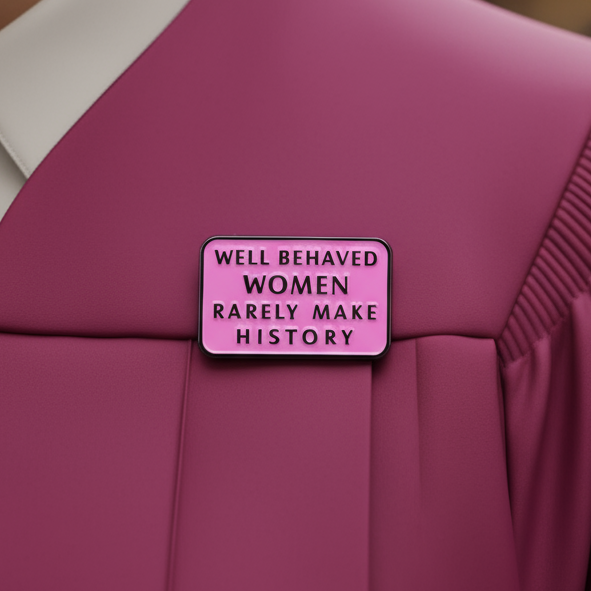Close-up of a pink enamel pin reading “Well Behaved Women Rarely Make History” on a pink jacket