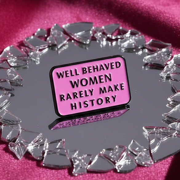 The Well Behaved Women Rarely Make History enamel pin, pink with bold black text, is centered on a shattered-mirror, fuchsia satin backdrop, symbolizing defiant empowerment and prideful self-expression.